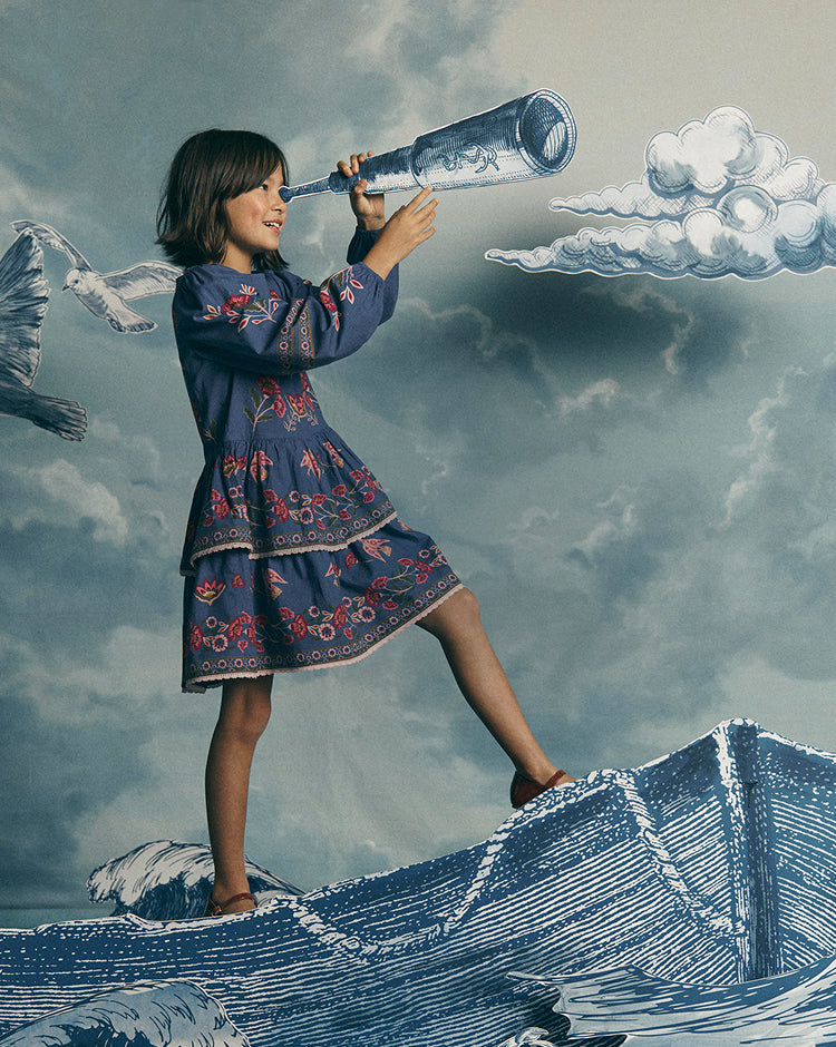 A young girl in a blue embroidered dress stands on illustrated waves looking through a large glass telescope against a cloudy sky backdrop.