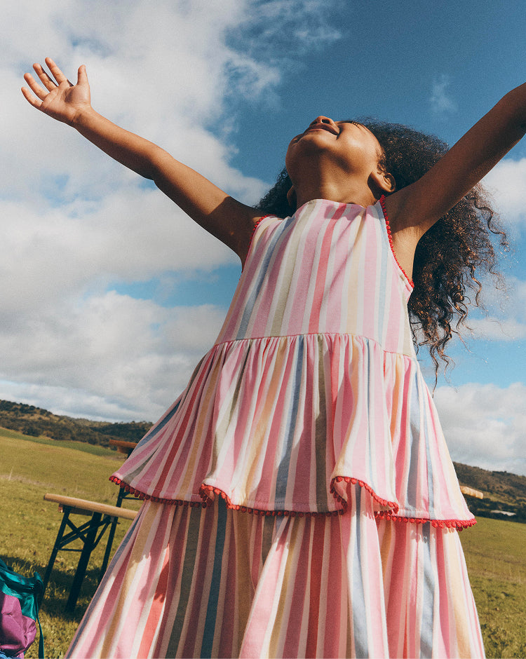 Girl in a striped dress with arms outstretched against a blue sky with clouds.