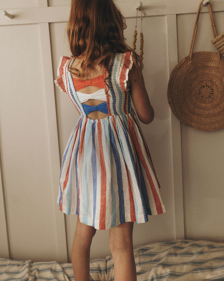 Girl wearing a colorful striped dress with a cutout back, standing against a wooden panel wall.