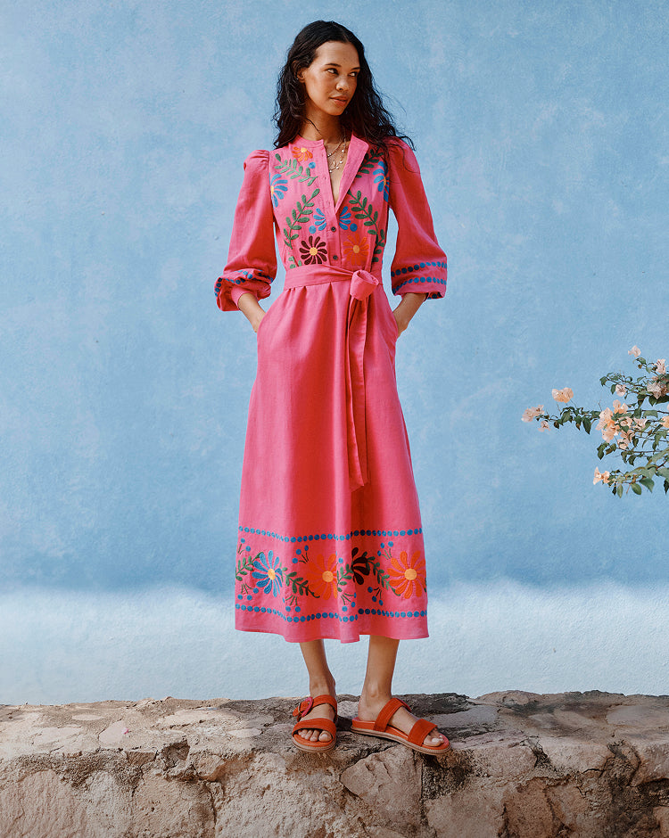 Woman wearing a pink dress with floral embroidery standing on a rock with a blue sky background
