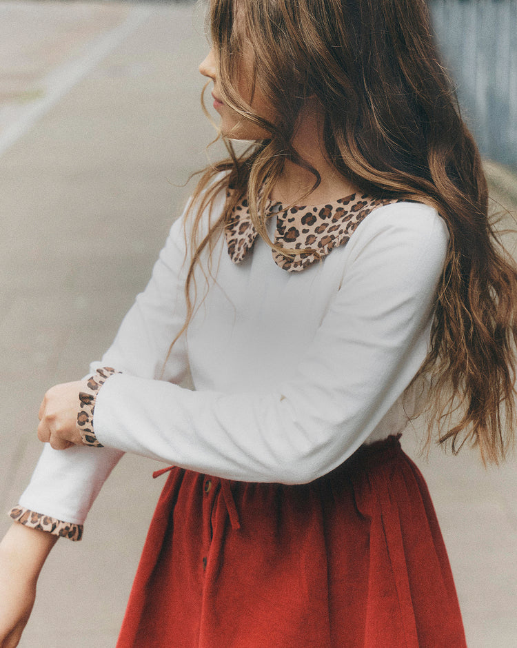 A young girl wearing a white long-sleeve top with a brown and beige leopard-print collar and matching leopard-print cuff trim, paired with a rust-red skirt, photographed outdoors.