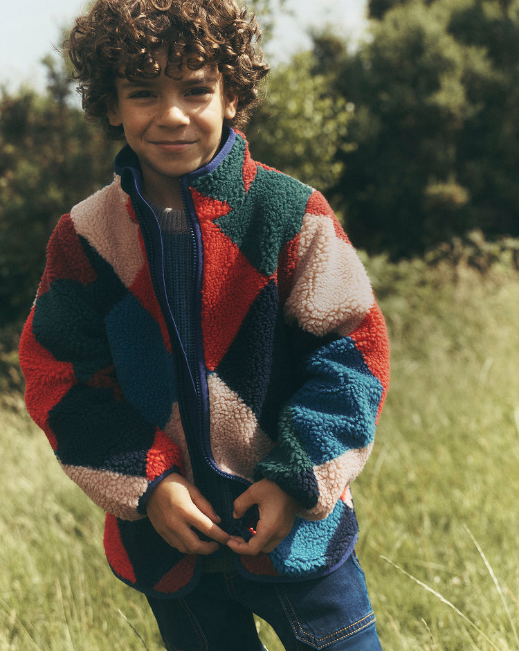 Child standing in a sunlit grassy field wearing a plush geometric fleece jacket in red, dark green, navy, blue and cream over a knitted jumper and blue jeans. The jacket has a high collar, front zip and contrasting colour-block pattern, with leafy trees softly blurred in the background.
