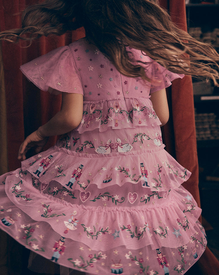 A young girl viewed from behind wearing a pink tiered tulle dress with embroidered fairy and nutcracker motifs, set against a dark, warm-toned background.
