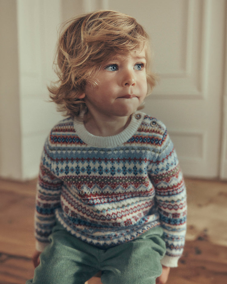 A toddler with wavy blond hair wearing a colorful Fair Isle patterned knit sweater and green corduroy pants, sitting on a wooden floor in front of a white paneled door.