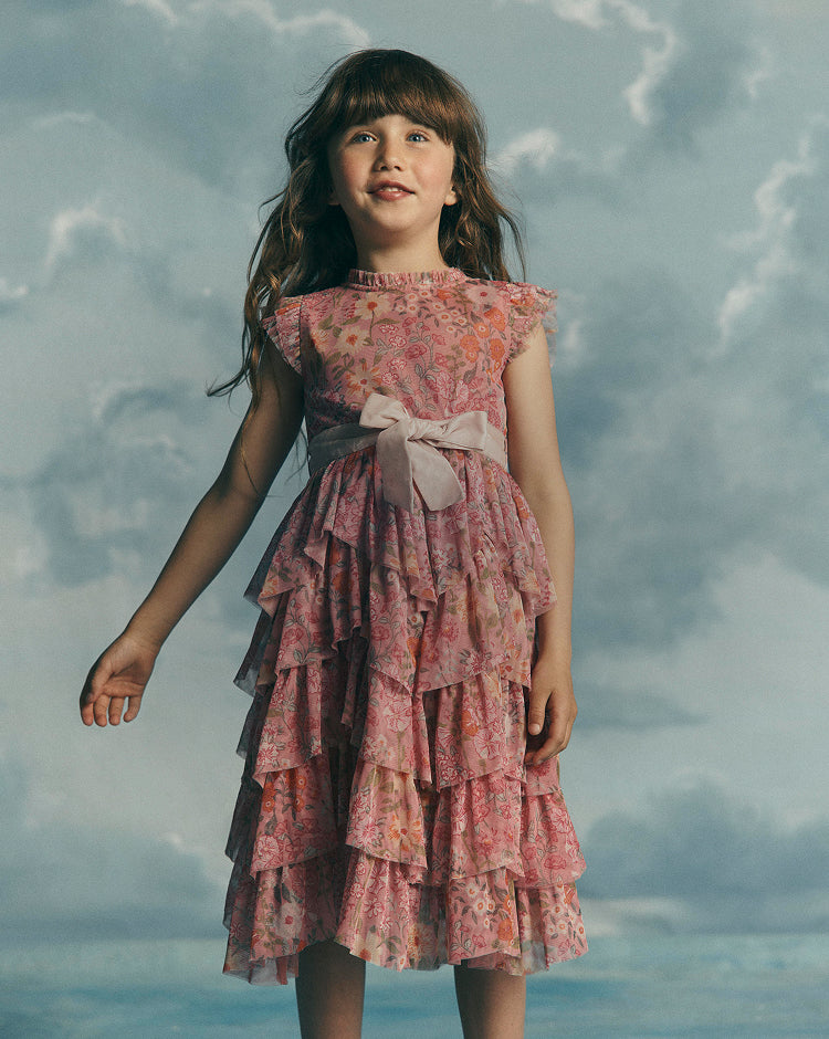 A young girl with long brown hair stands against a cloudy sky backdrop wearing a sleeveless pink floral tiered dress with a large bow at the waist.