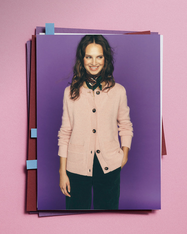 A fashion print or poster featuring a smiling woman in a light pink cardigan and dark trousers, set against a purple background and displayed on top of stacked papers against a pink wall.