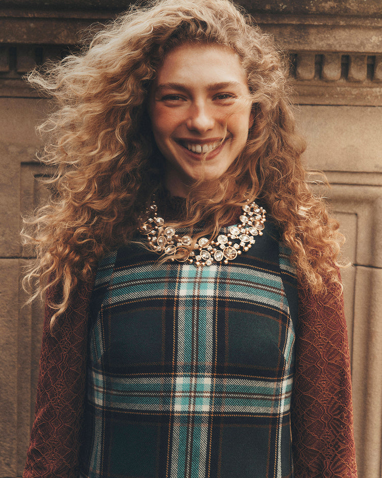 A smiling woman with voluminous curly light brown hair wears a dark teal and brown plaid top with a rust-colored textured cardigan and a large floral statement necklace, posed against a warm stone wall.