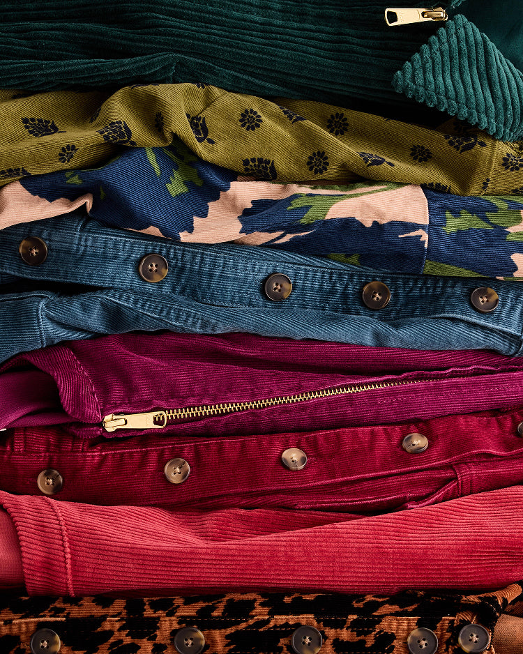 A close-up stack of folded garments in multiple colors and textures, including teal corduroy, olive green patterned fabric, blue floral print, denim, magenta corduroy, and pink fabric, with visible metal buttons and zippers.
