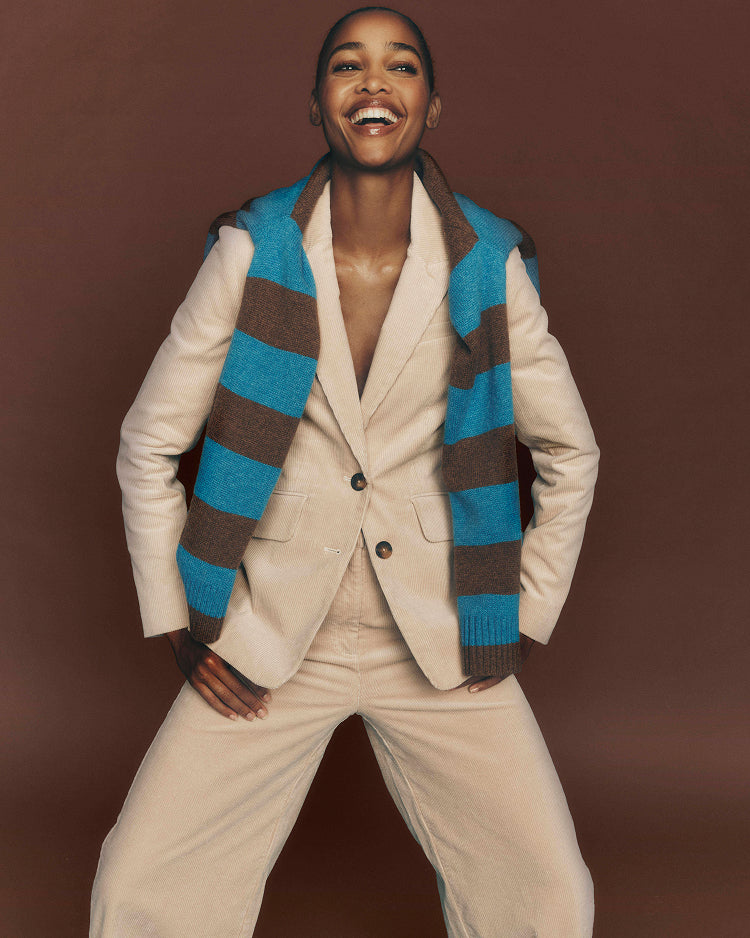 A smiling woman poses in a beige tailored blazer and matching trousers, layered with a blue and brown striped knit sweater vest, against a solid dark brown studio background.