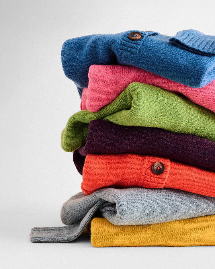 A stack of folded knit cardigans in multiple colors, including blue, pink, green, purple, orange, light gray, and mustard yellow, arranged on a light gray background.