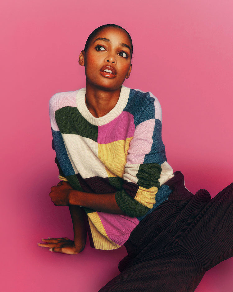 A model wearing a colorful patchwork knit sweater with blocks of pink, blue, green, yellow, and white, posed against a solid pink background.