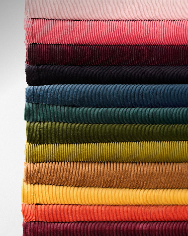 A vertical stack of folded corduroy fabric pieces in multiple colors, including pale pink, coral, burgundy, navy, teal, green, olive, mustard, tan, yellow, orange, and deep red, shown against a light neutral background.