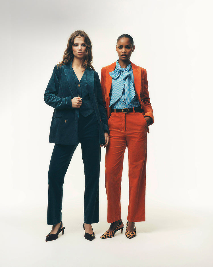 Two models standing against a plain white studio background. The model on the left wears a dark teal velvet pantsuit with black heels.
