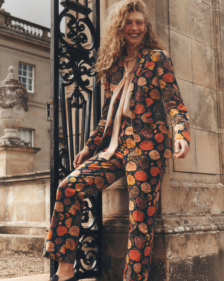 A woman with curly blonde hair models a matching two-piece suit set featuring a bold orange, red, and yellow floral pattern on a black background, styled with a cream-colored tie blouse, leaning against a stone wall next to an ornate black metal gate.