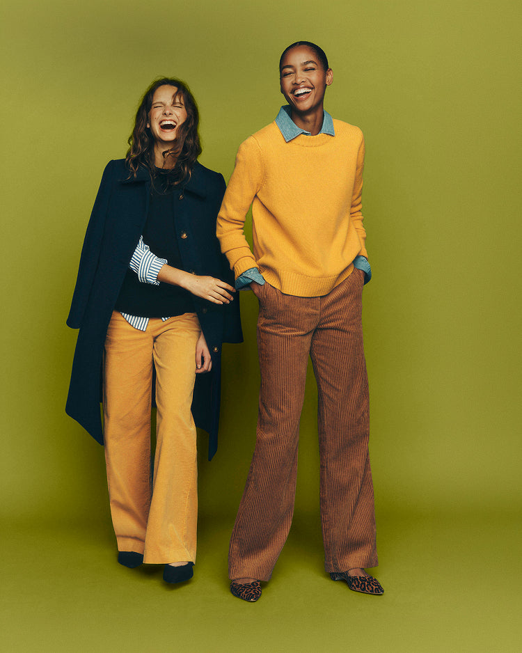 Two models posing together against a solid olive green studio backdrop. The model on the left wears a dark navy coat over a black top and mustard yellow flared trousers.