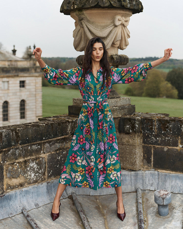 A woman stands outdoors on a stone terrace wearing a long-sleeved midi dress with a colorful floral print on a teal background. She has her arms outstretched and is posing in front of a stone urn and a large historic building.