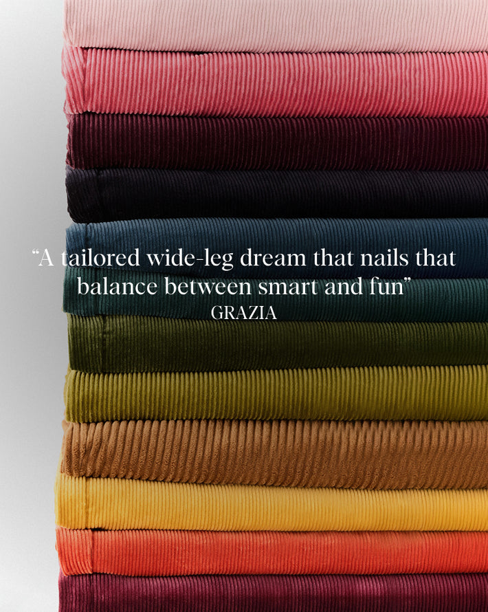 A vertical stack of folded corduroy fabric in multiple colors, including pink, burgundy, navy, teal, olive green, mustard yellow, orange, and deep red. White text overlaid in the center reads, "A tailored wide-leg dream that nails that balance between smart and fun" GRAZIA.