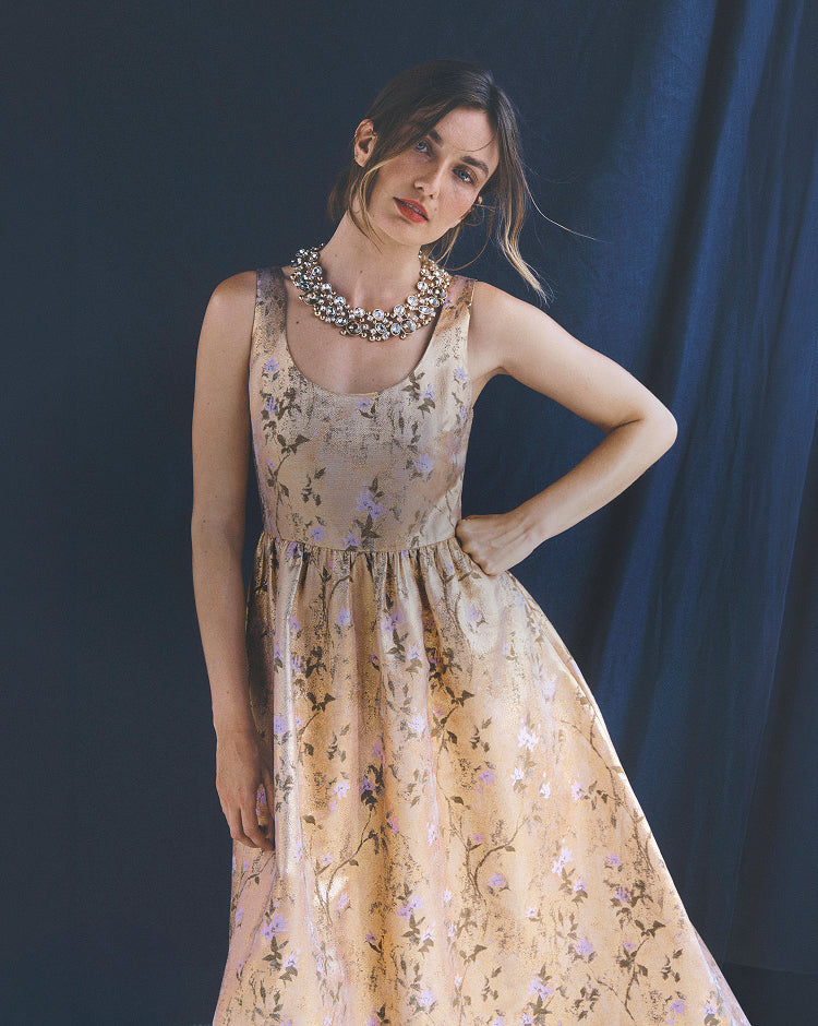A woman models a sleeveless beige A-line dress with a delicate purple and green floral pattern, styled with a chunky silver statement necklace against a dark blue studio backdrop.