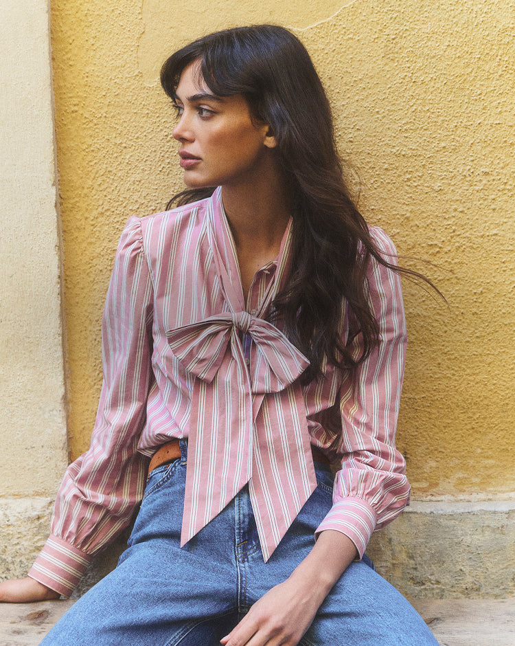 Woman wearing a pink striped blouse with a bow tie, sitting against a yellow wall.