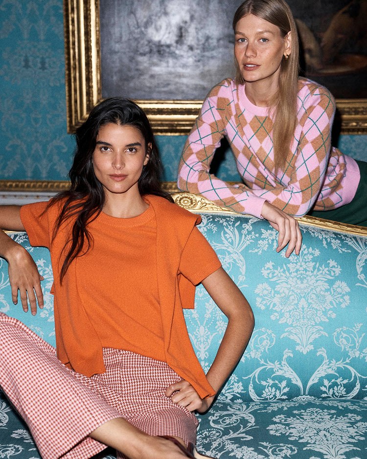Two models posing on a teal damask sofa in front of a gold-framed painting. The model in the foreground wears an orange short-sleeve top and pink-and-white gingham trousers, while the model in the background wears a pink argyle sweater.