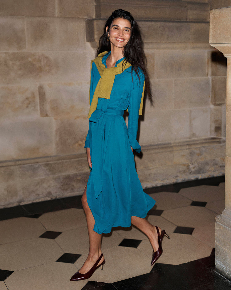 A woman wearing a teal blue midi dress with a yellow collar detail and matching belt, walking in an indoor stone hallway.