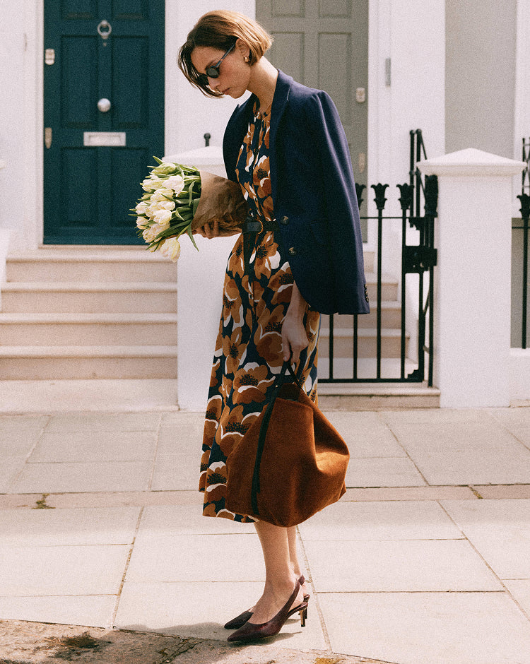 Woman in a patterned dress and navy coat holding flowers, standing on a sidewalk.