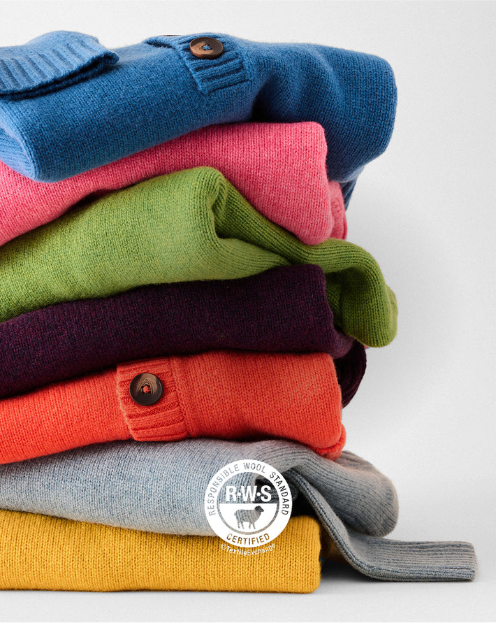 A stack of folded knit cardigans in multiple colors, including blue, pink, green, purple, orange, gray, and mustard yellow, photographed on a light gray studio background. A circular RWS Certified logo is overlaid near the bottom.