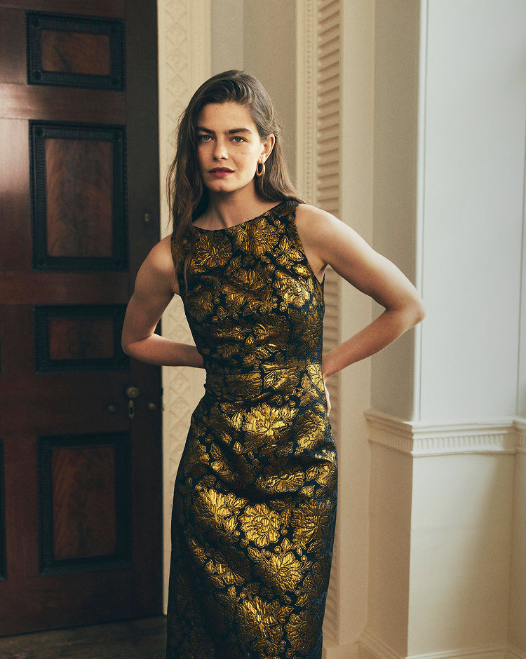 A woman stands indoors wearing a sleeveless, floor-length black dress with an ornate metallic gold floral or leaf-like pattern. The photo is taken in a real interior setting with a dark wooden door on the left and cream-colored wall molding on the right.