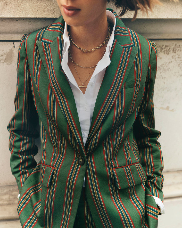 A close-up of a person wearing a green tailored blazer with vertical red and orange stripes, layered over a white collared shirt and gold necklaces, standing against a light stone wall.