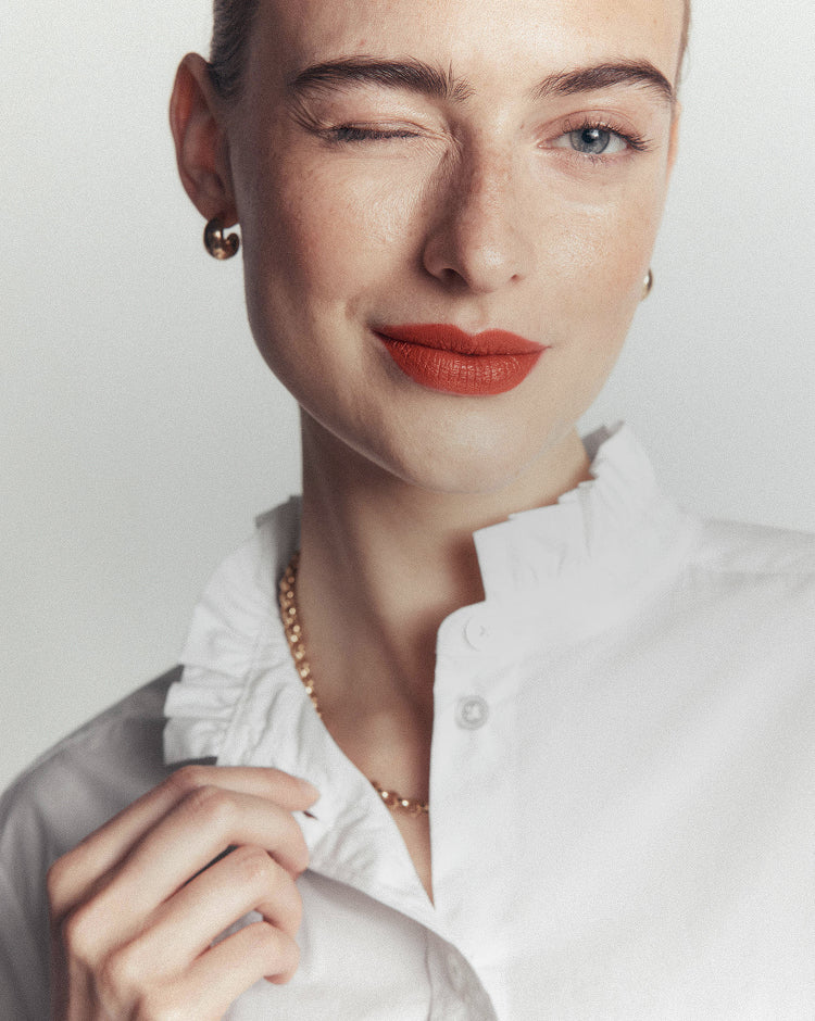 Close-up portrait of a woman wearing a white ruffled collar blouse, gold chain necklace, and small gold hoop earrings against a light gray background.