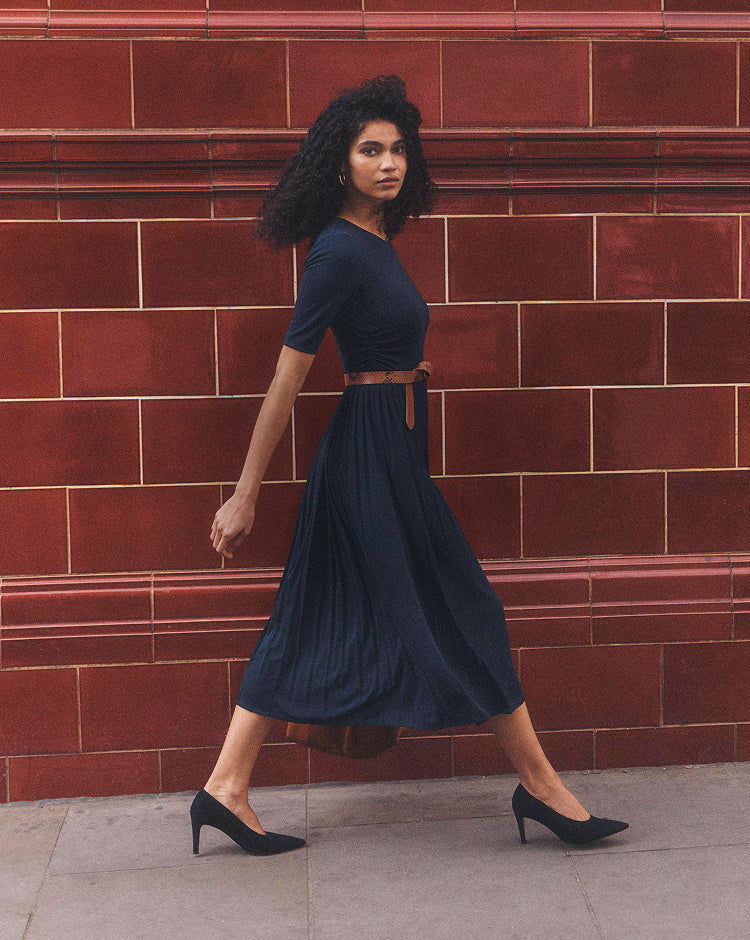 A woman wearing a navy blue midi dress with a brown belt walks in front of a red brick wall.