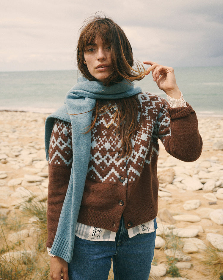 A woman stands on a rocky beach wearing a brown Fair Isle cardigan with a white geometric pattern, a light blue knit scarf, and blue jeans.