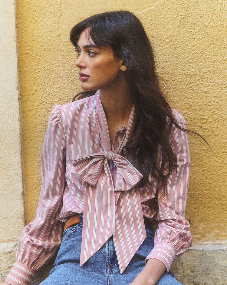 Woman wearing a pink and white striped blouse with a bow tie, sitting against a yellow wall.