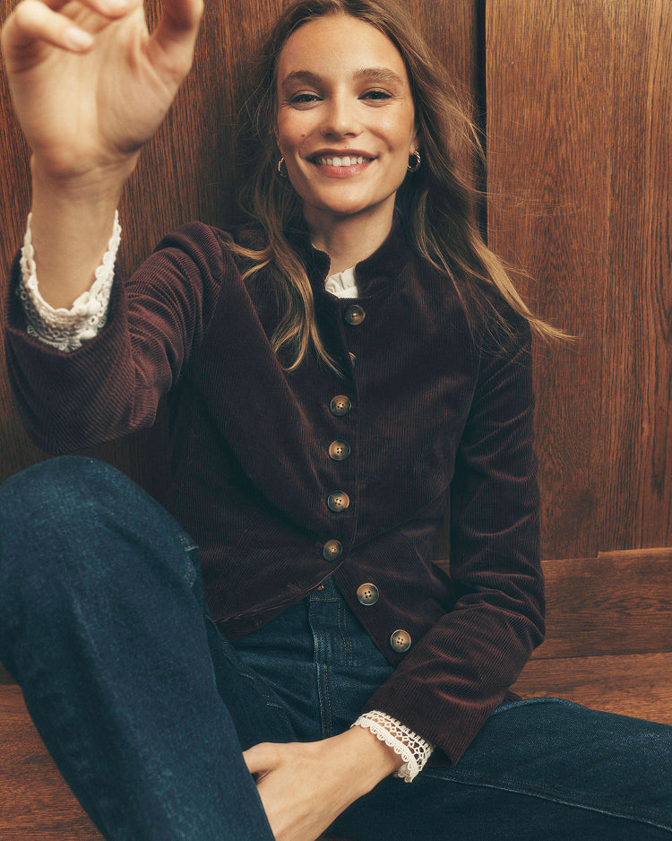A woman wearing a dark brown corduroy jacket with gold buttons and white lace cuffs, paired with dark blue jeans, sitting against a wooden wall.
