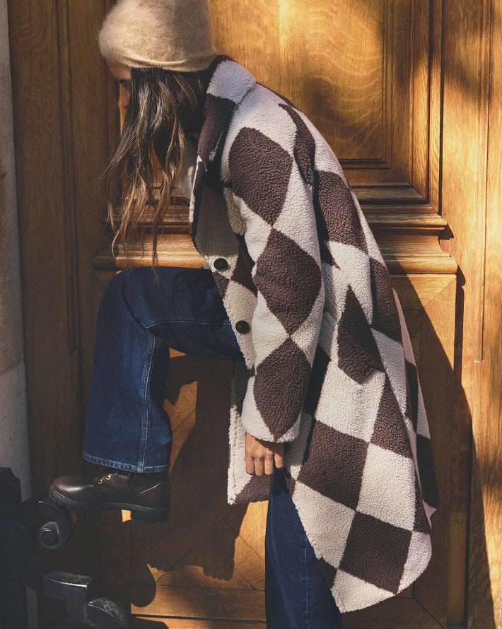 A person wearing a brown and cream argyle sherpa coat, blue jeans, and a beige beanie sits on a wooden bench in warm sunlight.