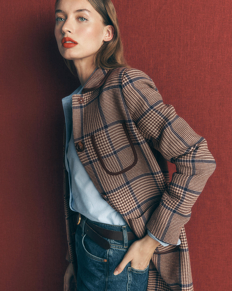 A woman wearing a brown and beige plaid blazer over a light blue shirt and blue jeans, posing against a textured dark red wall.