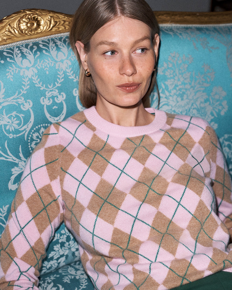 A woman wearing a pink and tan argyle sweater with thin green lines, seated on a teal damask armchair with a gold armrest.