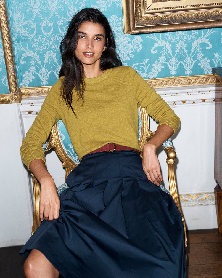 A woman seated in a gold armchair wearing a mustard yellow long-sleeve knit top and a dark navy skirt, posed against a blue damask wallpaper and ornate gold-framed wall decor.