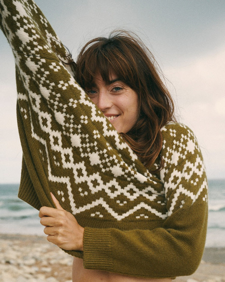A woman on a beach wearing a ribbed knit sweater with a brown and off-white geometric pattern, holding the sleeve up in a pose against a cloudy sky and ocean background.