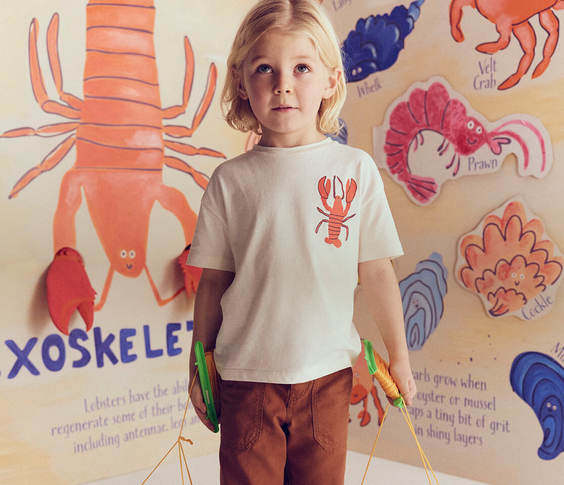 Child wearing a white t-shirt with a red lobster design, standing in front of a wall with colorful illustrations of sea creatures.