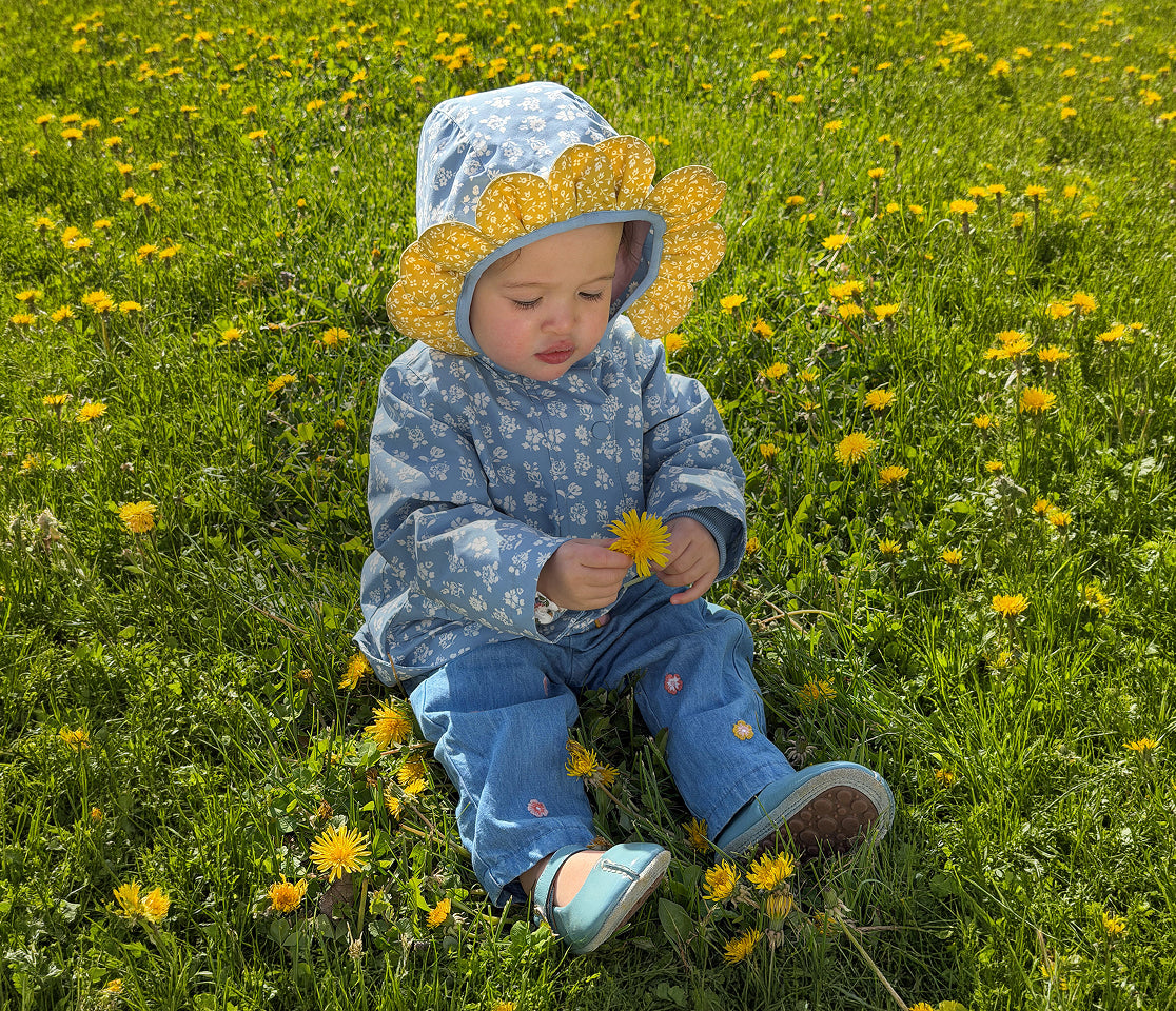 Child in a floral hat and blue coat sitting in a field of yellow flowers.