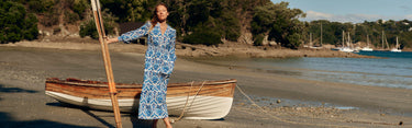 A woman in a blue dress by Boden stands next to a boat, with a cheerful expression in a scenic outdoor location.
