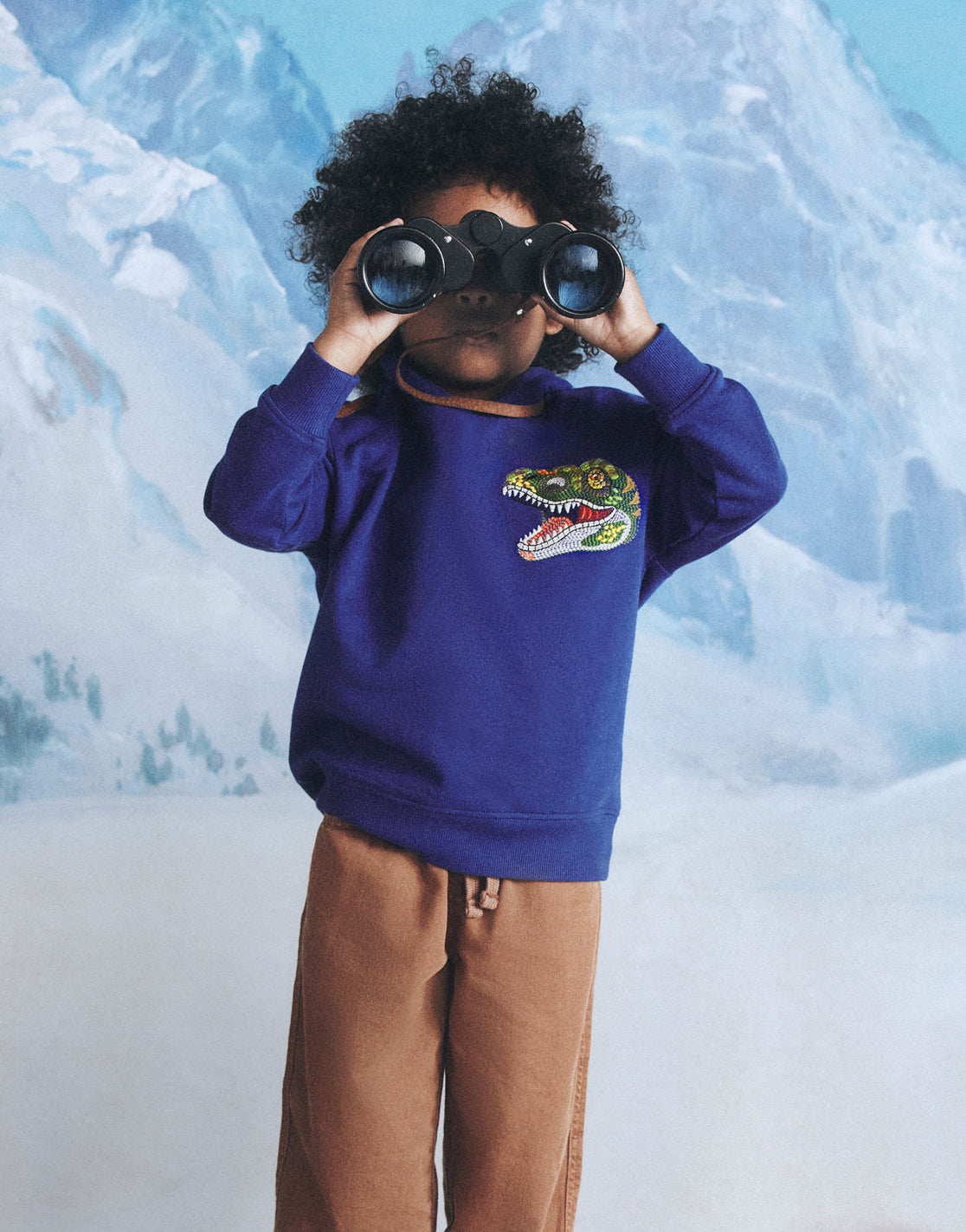 Child wearing a blue sweatshirt with a colorful logo, standing against a snowy mountain background.