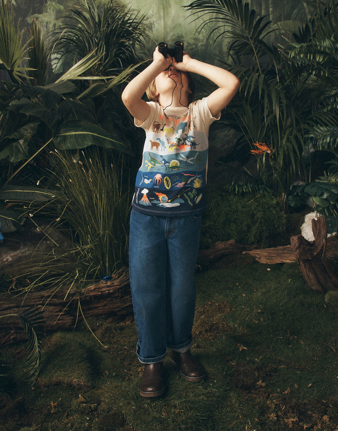 Person standing in a forest with binoculars, wearing a colorful shirt and blue jeans.