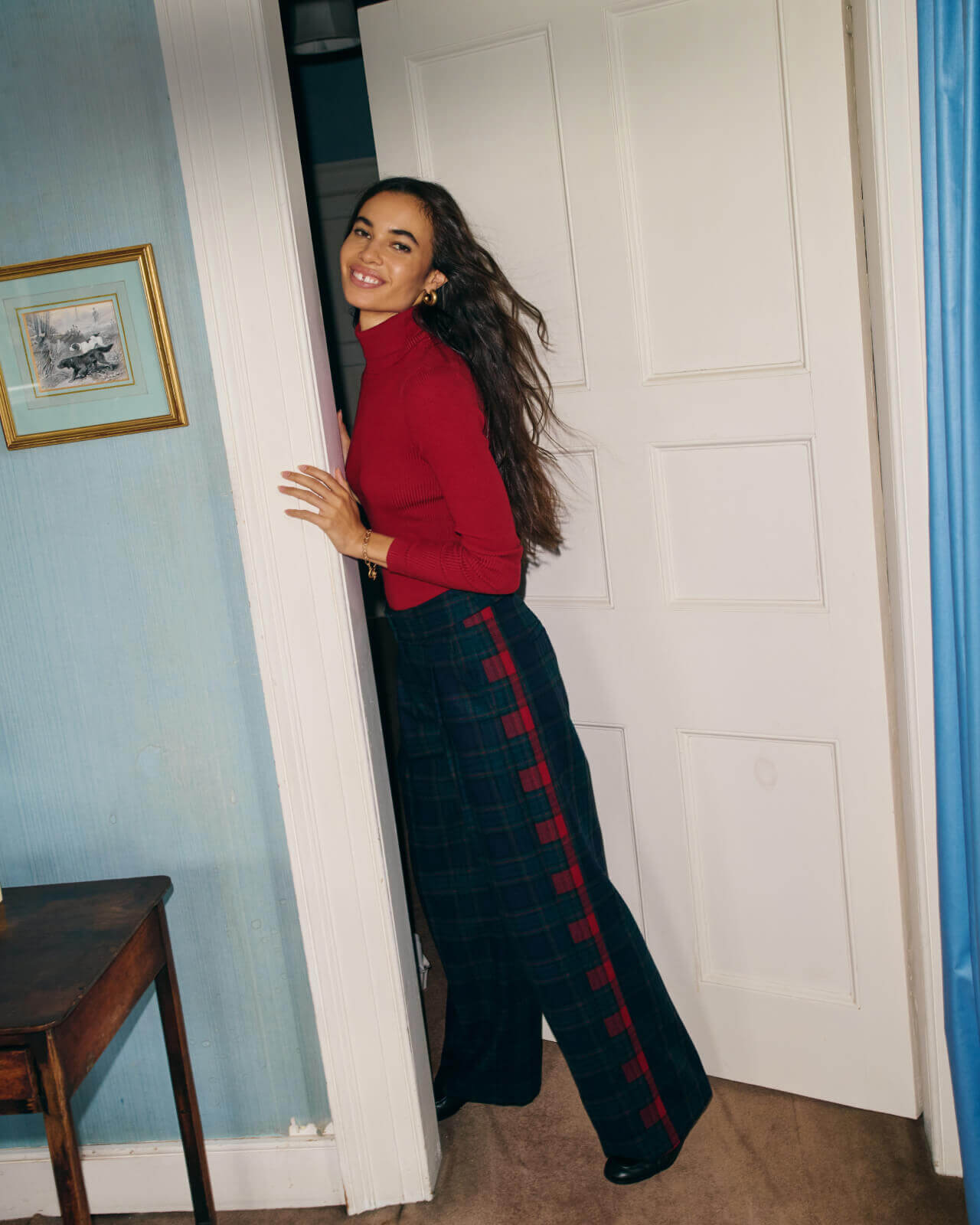 Woman standing in the doorway wearing a red polo neck top and green plaid trousers. 