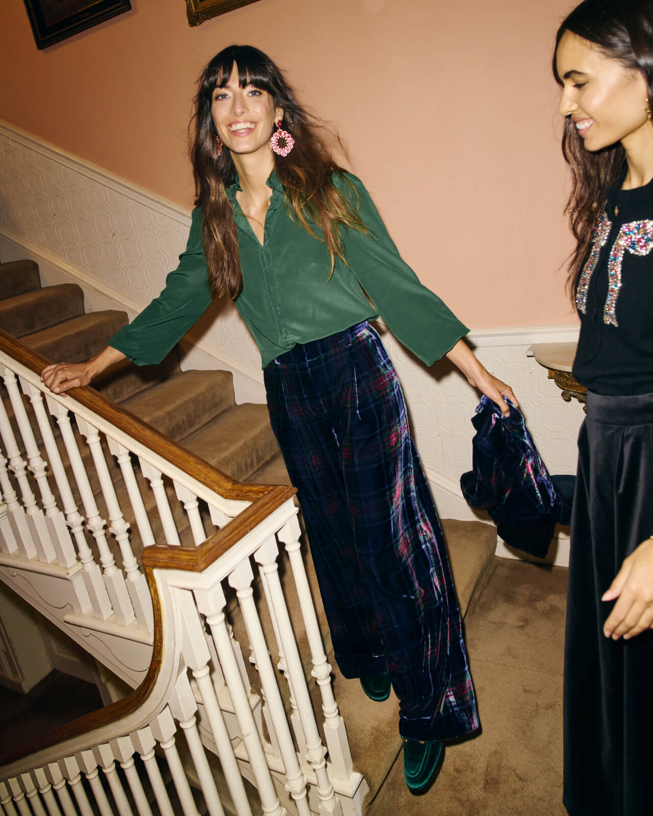 Woman with a green shirt and purple plaid trousers standing on the stairs holding a matching plaid jacket.