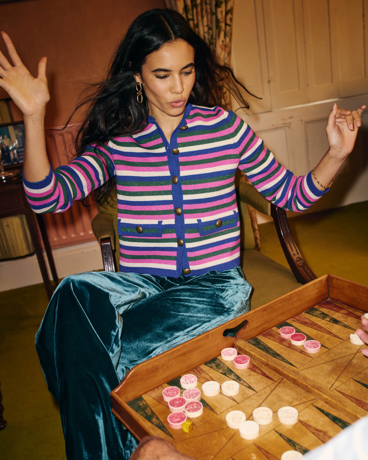 Woman wearing a striped cardigan and green velvet pants playing a board game. 